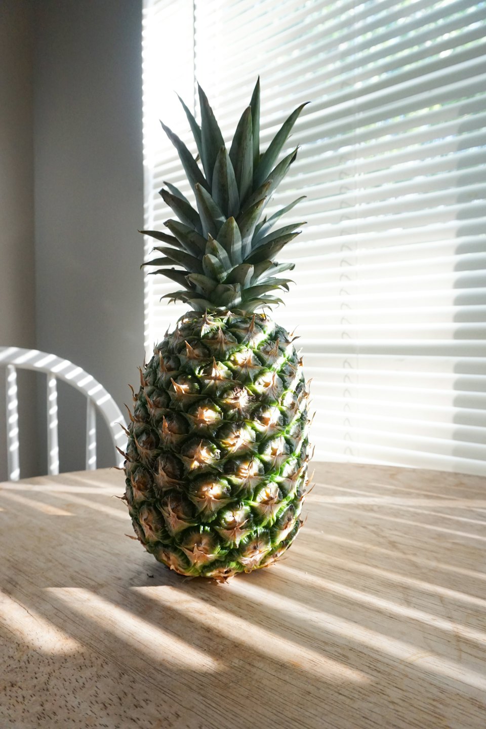 A fresh pineapple stands on a wooden table with striped shadows.